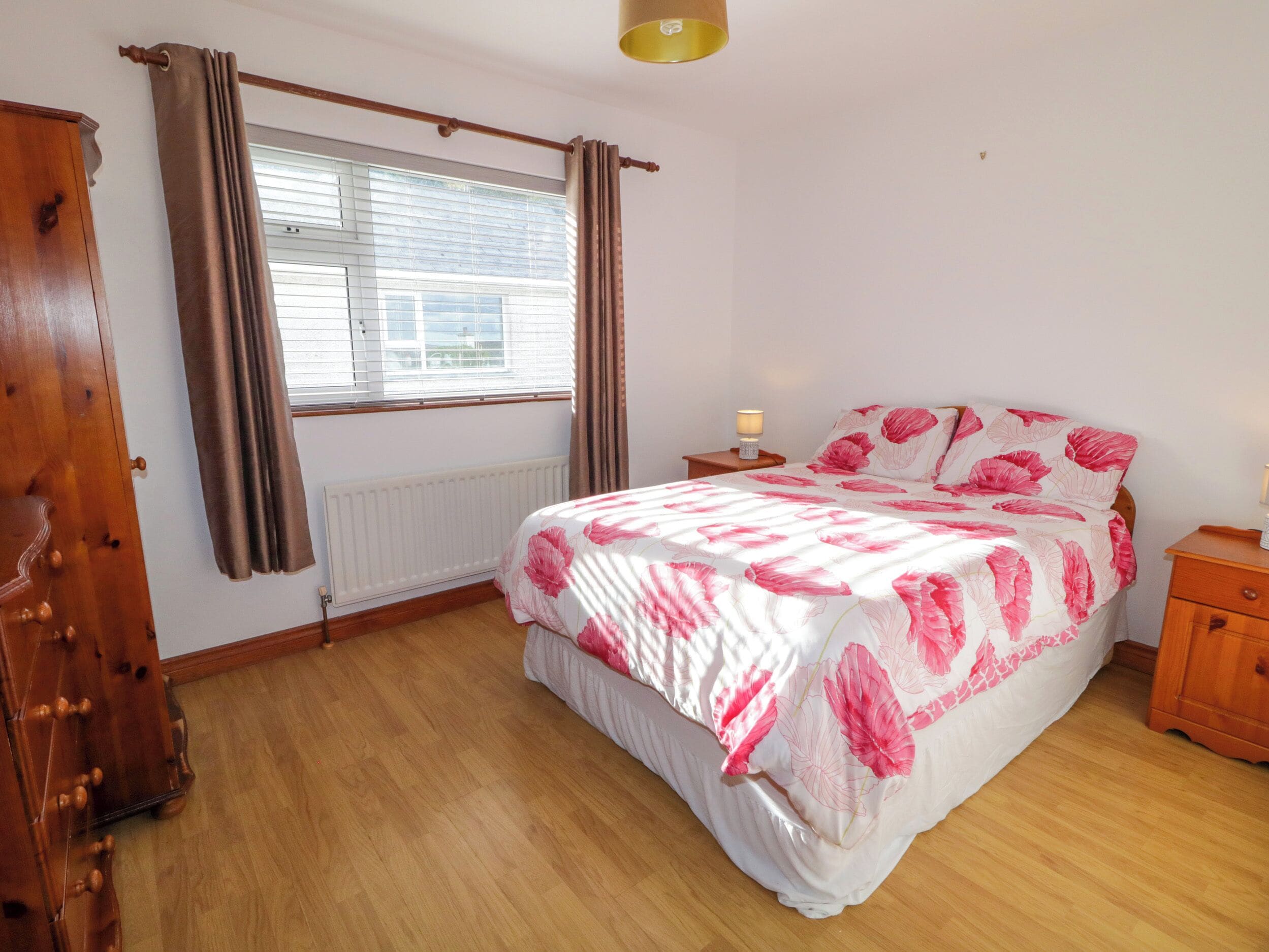 A bedroom with a bed, wooden floor, window with curtains, bedside table with lamp, and a radiator.