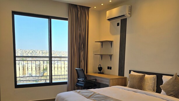 Ein Hotelzimmer mit einem großen Bett, einem Schreibtisch, einem Sessel, einer Klimaanlage und Blick auf die Stadt durch ein großes Fenster.