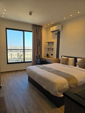 Habitación de hotel con una cama grande, un escritorio, una silla, un equipo de aire acondicionado y vistas a la ciudad a través de un ventanal amplio.