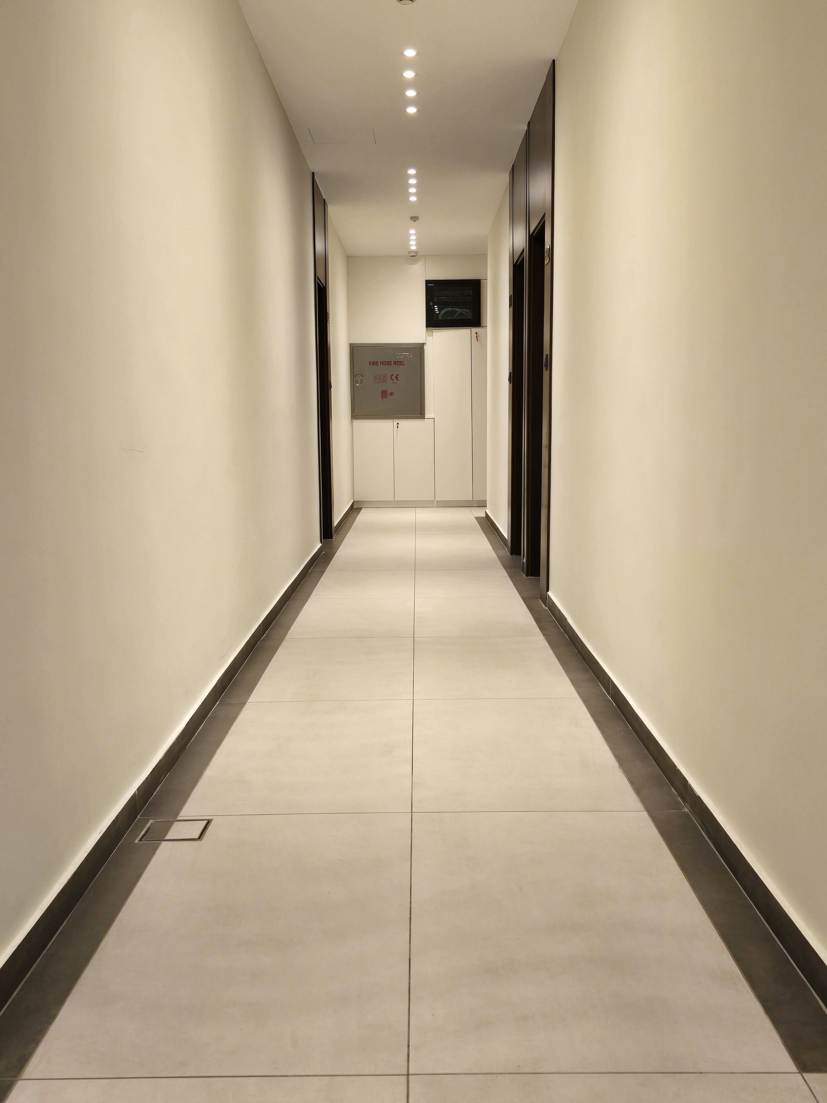 Un long couloir étroit, doté d’un sol carrelé et de murs blancs. Des portes se trouvent de part et d’autre, et un panneau sur le mur indique l’emplacement d’un enrouleur de tuyau d’incendie.