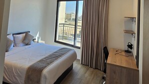 Ein modernes Hotelzimmer mit einem großen Bett, einem Schreibtisch mit Stuhl und Blick auf die Stadt durch ein großes Fenster.