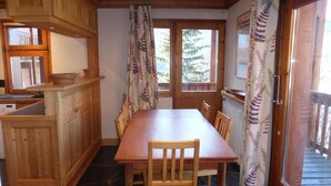 Une table à manger en bois avec des chaises, un coin cuisine avec des meubles en bois, et une vue sur l’extérieur par une porte.