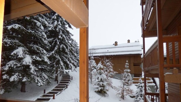 Un balcon en bois offrant une vue sur un paysage enneigé, avec des arbres et un bâtiment dont le toit est recouvert de neige.