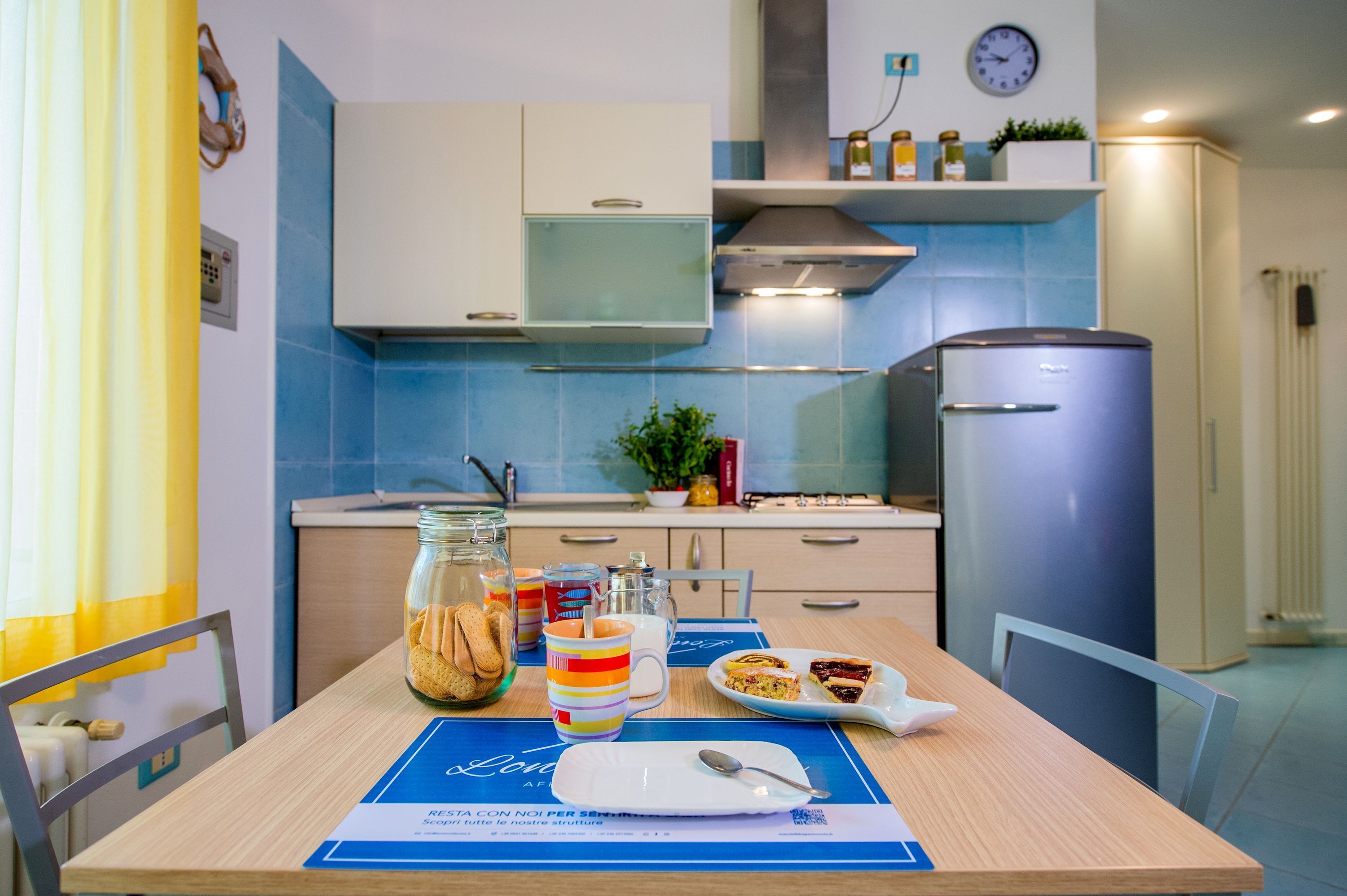 A modern kitchen with a blue tiled backsplash, a stainless steel refrigerator, and a dining table set with a plate of food, a mug, and a glass jar.