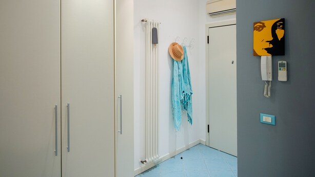 Un pasillo con un armario blanco, un radiador, suelo de baldosas azules y un teléfono fijado a la pared.