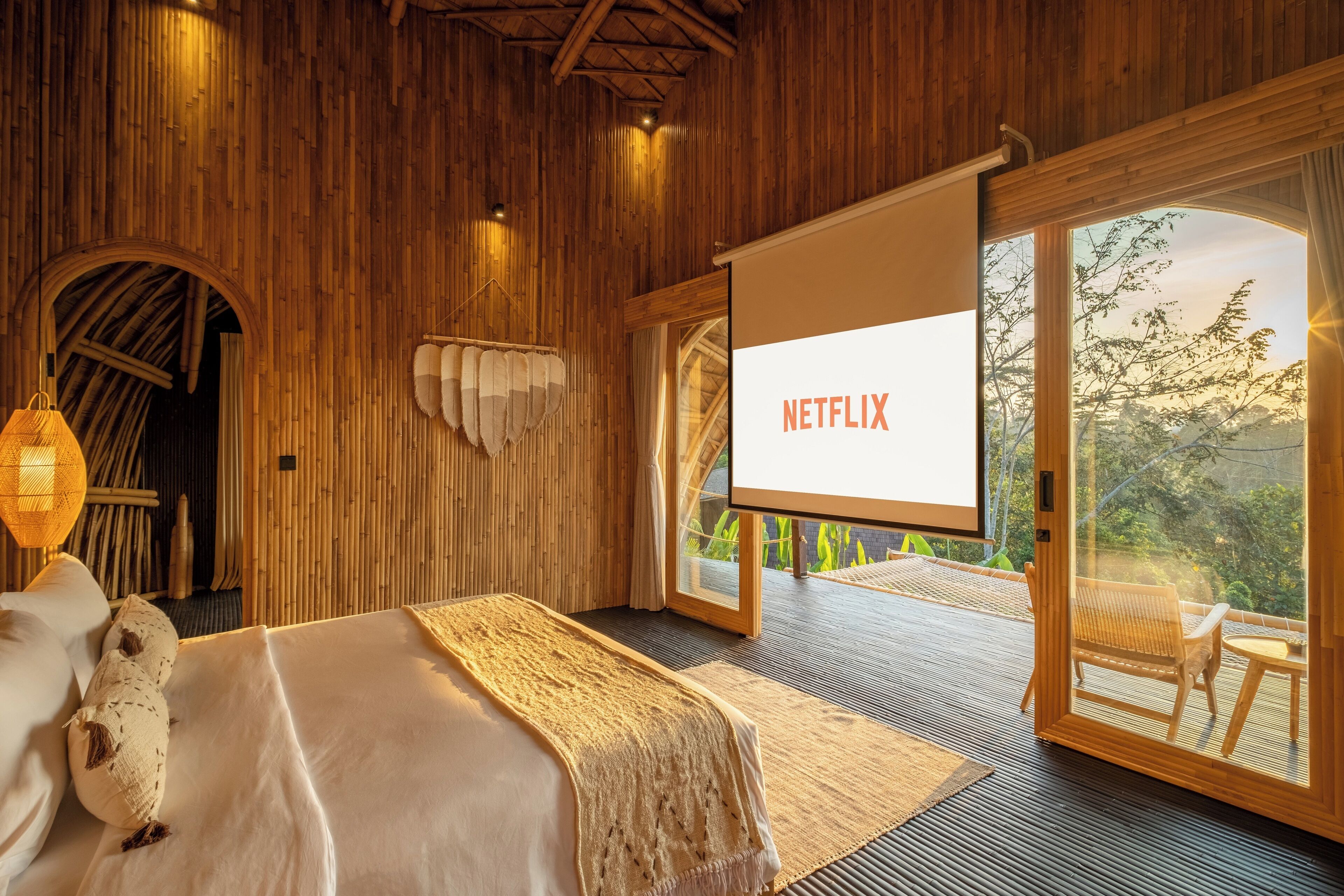 A room with a bed, a projector screen displaying 'NETFLIX', and a balcony with a view of trees.