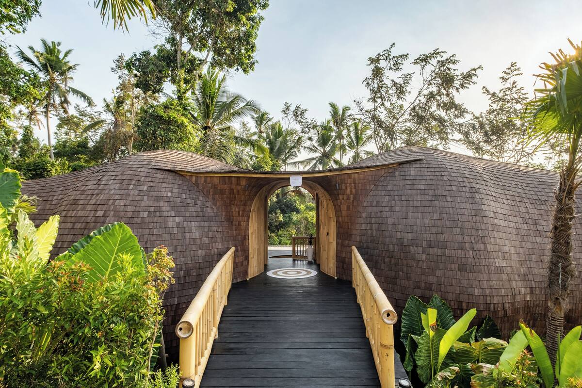A wooden bridge leading to a structure with a curved roof, surrounded by lush greenery and palm trees.
