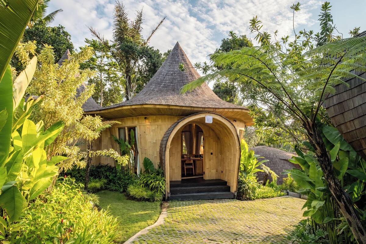 A wooden house with a conical roof, surrounded by lush greenery and a cobblestone path leading to the entrance.