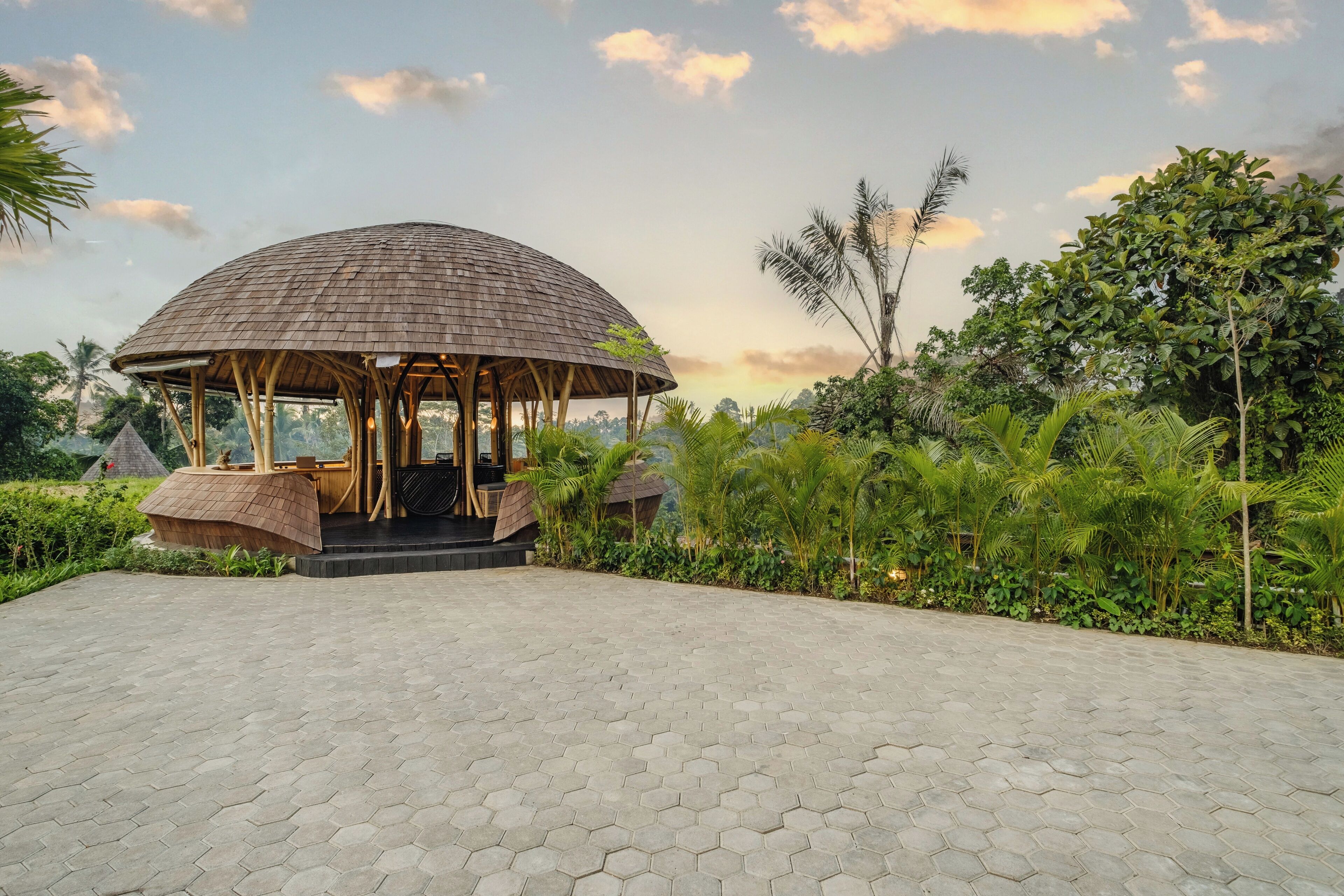 A hexagonal pavilion with a wooden roof and seating area, surrounded by lush greenery and a paved walkway.