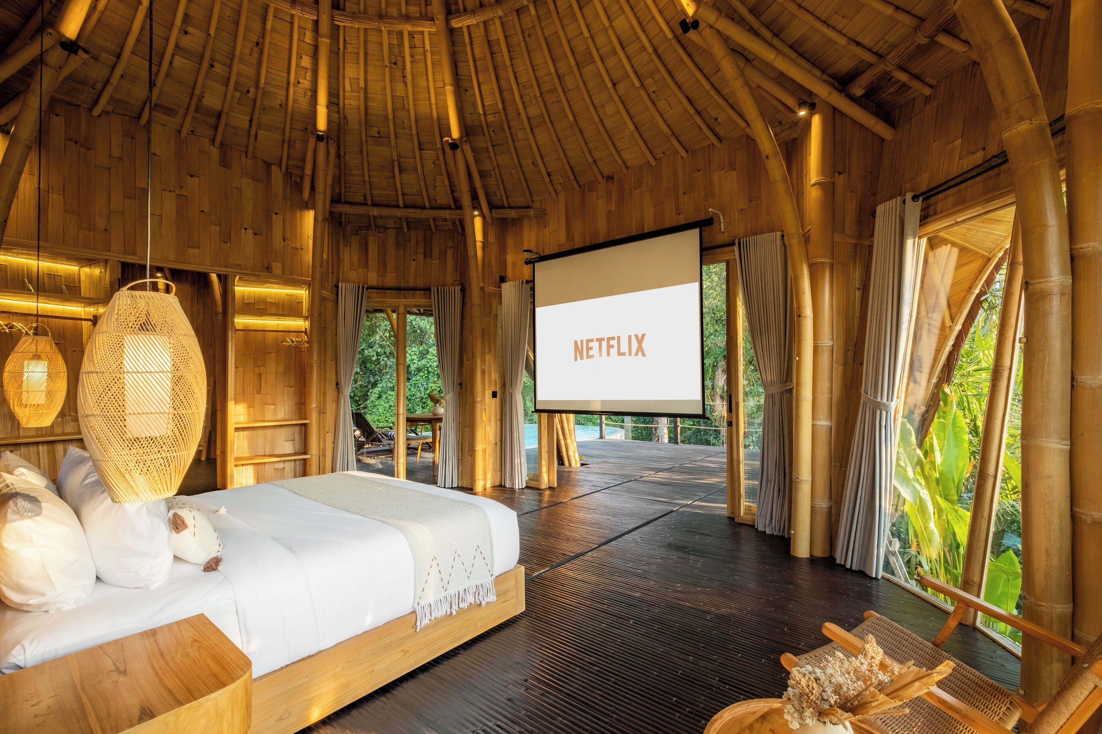 A room with a bed, a projector screen displaying 'NETFLIX', and a view of greenery outside.