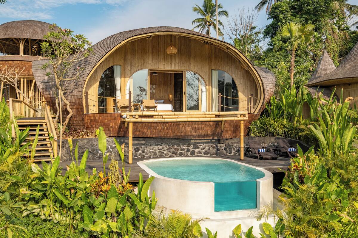A wooden villa with a curved roof, a pool, and lush tropical vegetation.