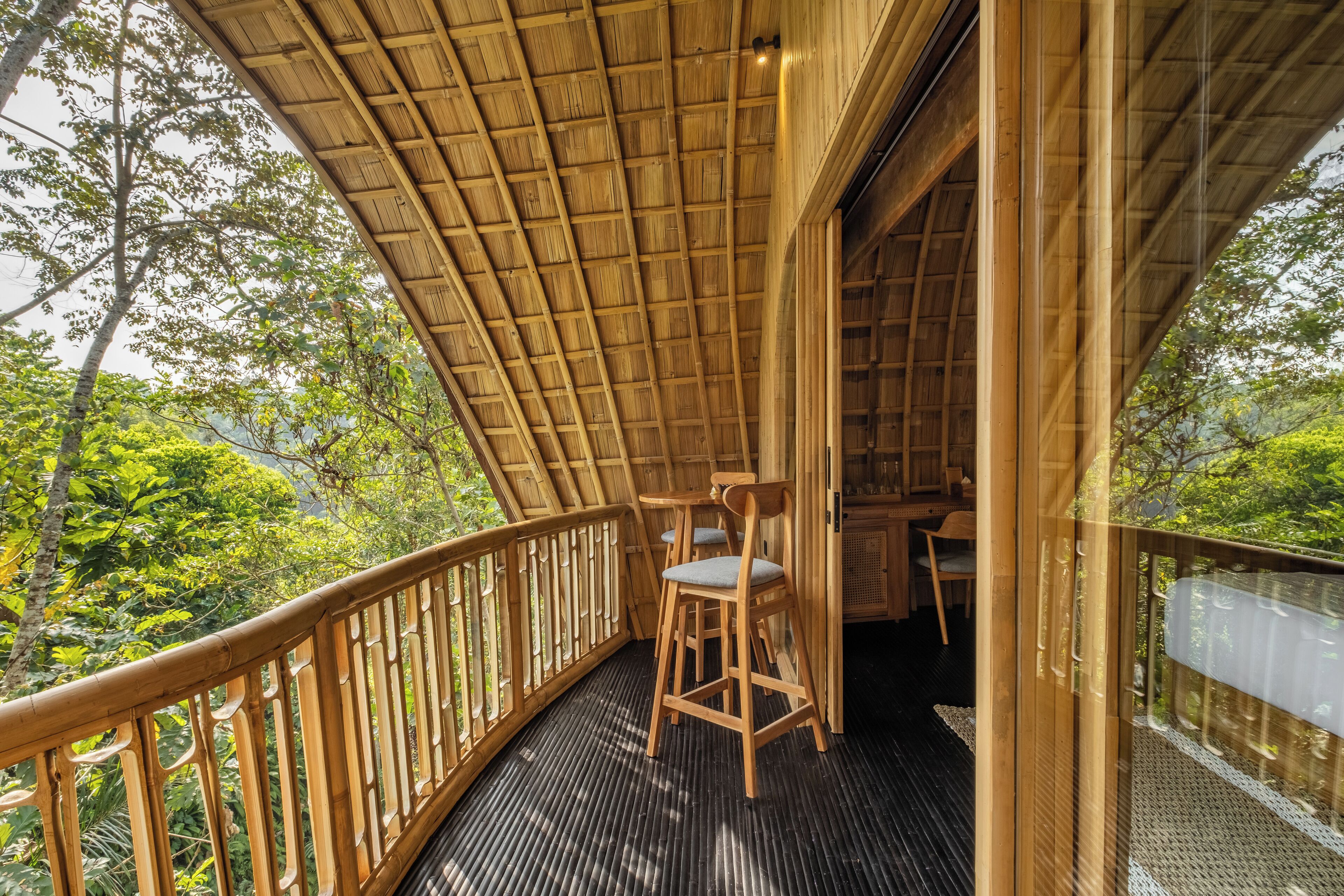 A wooden deck with a thatched roof, two wooden chairs, and a view of lush greenery.