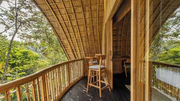 A wooden deck with a thatched roof, two wooden chairs, and a view of lush greenery.