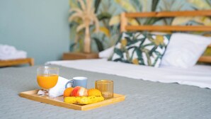Bandeja de desayuno con jugo de naranja, una taza, un plátano y una manzana sobre una bandeja de madera en una habitación de hotel con una cama y una pared con motivos tropicales.