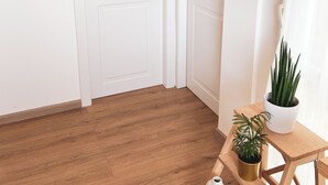 Una habitación con piso de madera, paredes blancas, una puerta cerrada con manija de latón y una pequeña mesa de madera con dos plantas en macetas.