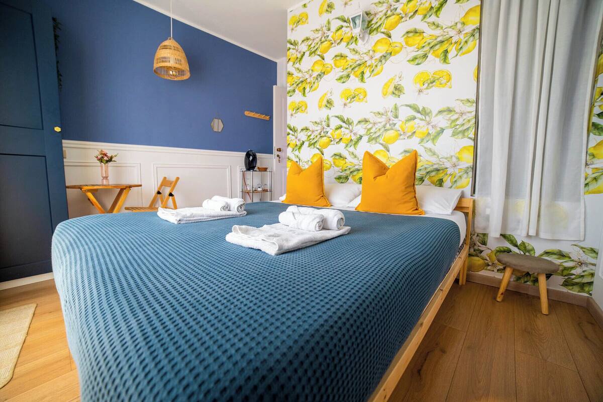 Una cama bien hecha con ropa de cama azul, dos almohadas amarillas y toallas blancas enrolladas.