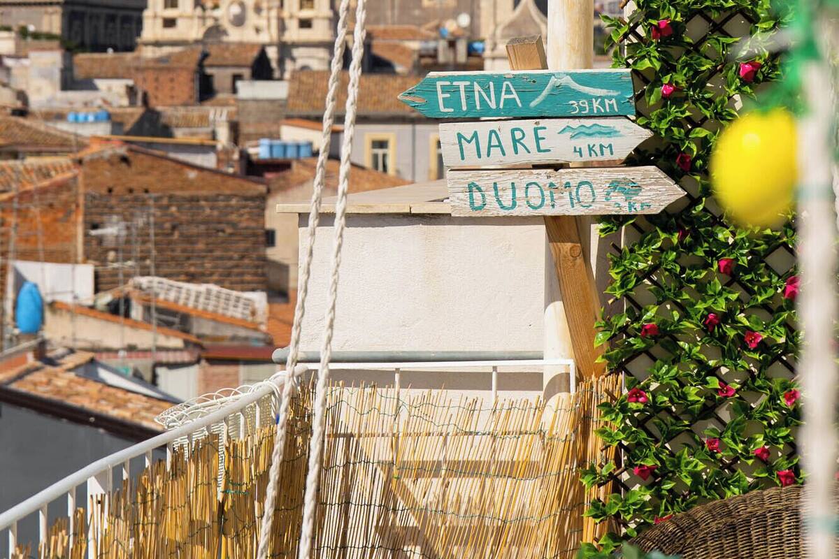 Una señal de calle indica direcciones hacia Etna, Mare y Duomo, con una montaña nevada y edificios históricos de fondo.