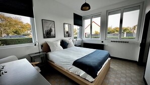 A modern bedroom with a bed, bedside tables, a desk, and large windows offering a view of the outside. - 700m from the sea, apartment with terrace, parking and pétanque court. (Cayeux-sur-Mer)