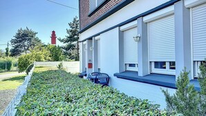 A two-story building with a brick facade and white trim. There are two windows with shutters, a small porch with a bench, and a red structure in the background. - 700m from the sea, apartment with terrace, parking and pétanque court. (Cayeux-sur-Mer)