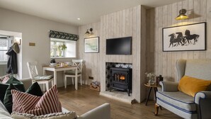 A living room with a fireplace, a dining area, and a TV mounted on the wall. - Fabulously presented dog friendly holiday cottage with outside tub - The Coach House, South Cerney (South Cerney)