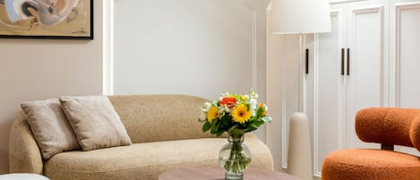 A living room with a beige sofa, a round wooden coffee table, a glass vase with flowers, a tall floor lamp, and a vibrant orange armchair.