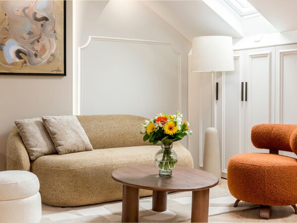 A living room with a beige sofa, a round wooden coffee table, a glass vase with flowers, a tall floor lamp, and a vibrant orange armchair. - Merveil - Luxury Suite - Champs Elysees - St Cyr I (Paris)
