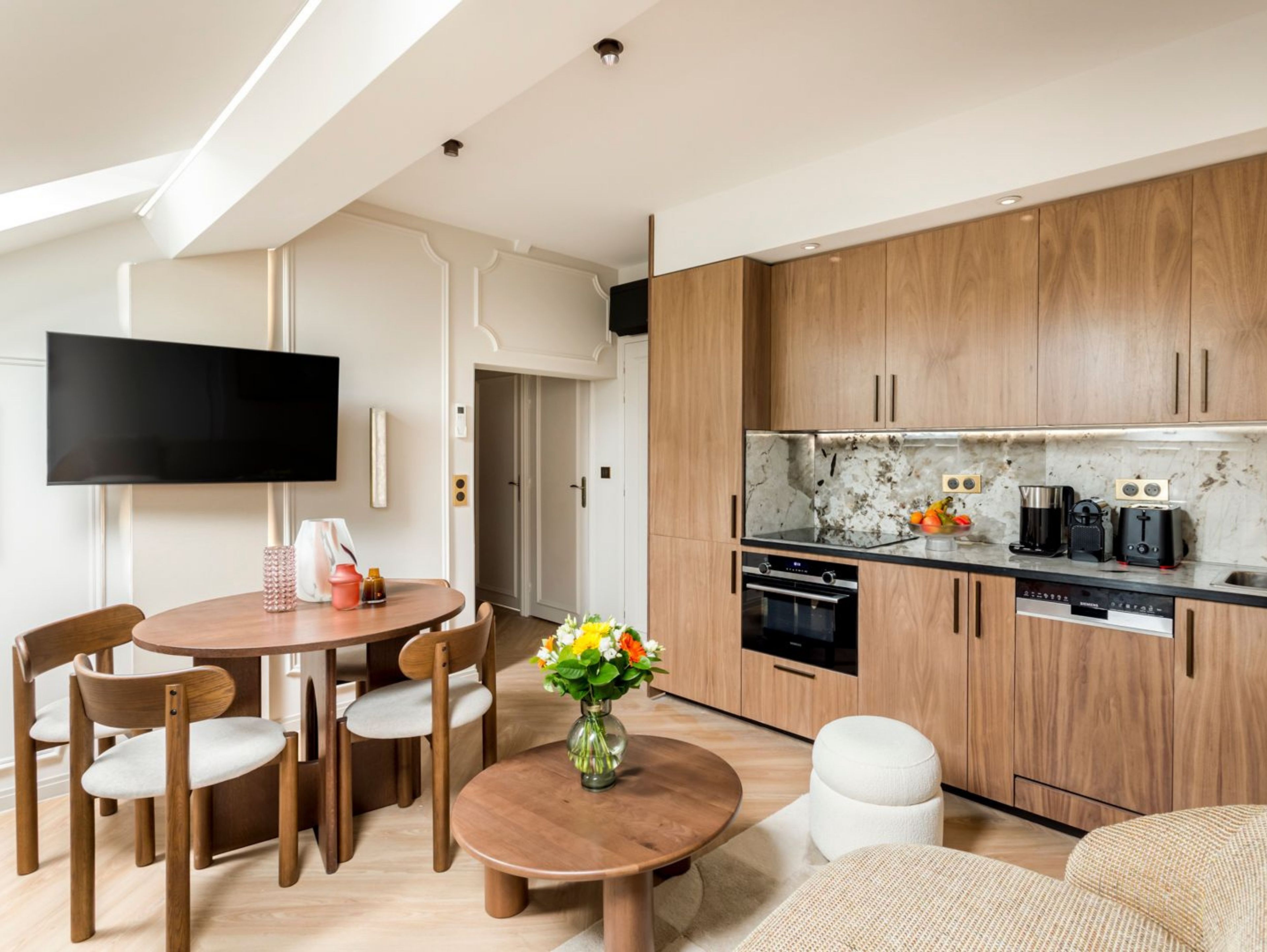 A modern kitchen with wooden cabinets, a black countertop, and a built-in oven. There is a dining area with a round table and chairs, and a flat-screen TV mounted on the wall.