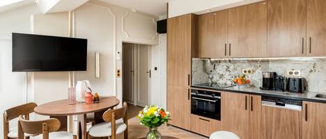 A modern kitchen with wooden cabinets, a black countertop, and a built-in oven. There is a dining area with a round table and chairs, and a flat-screen TV mounted on the wall.