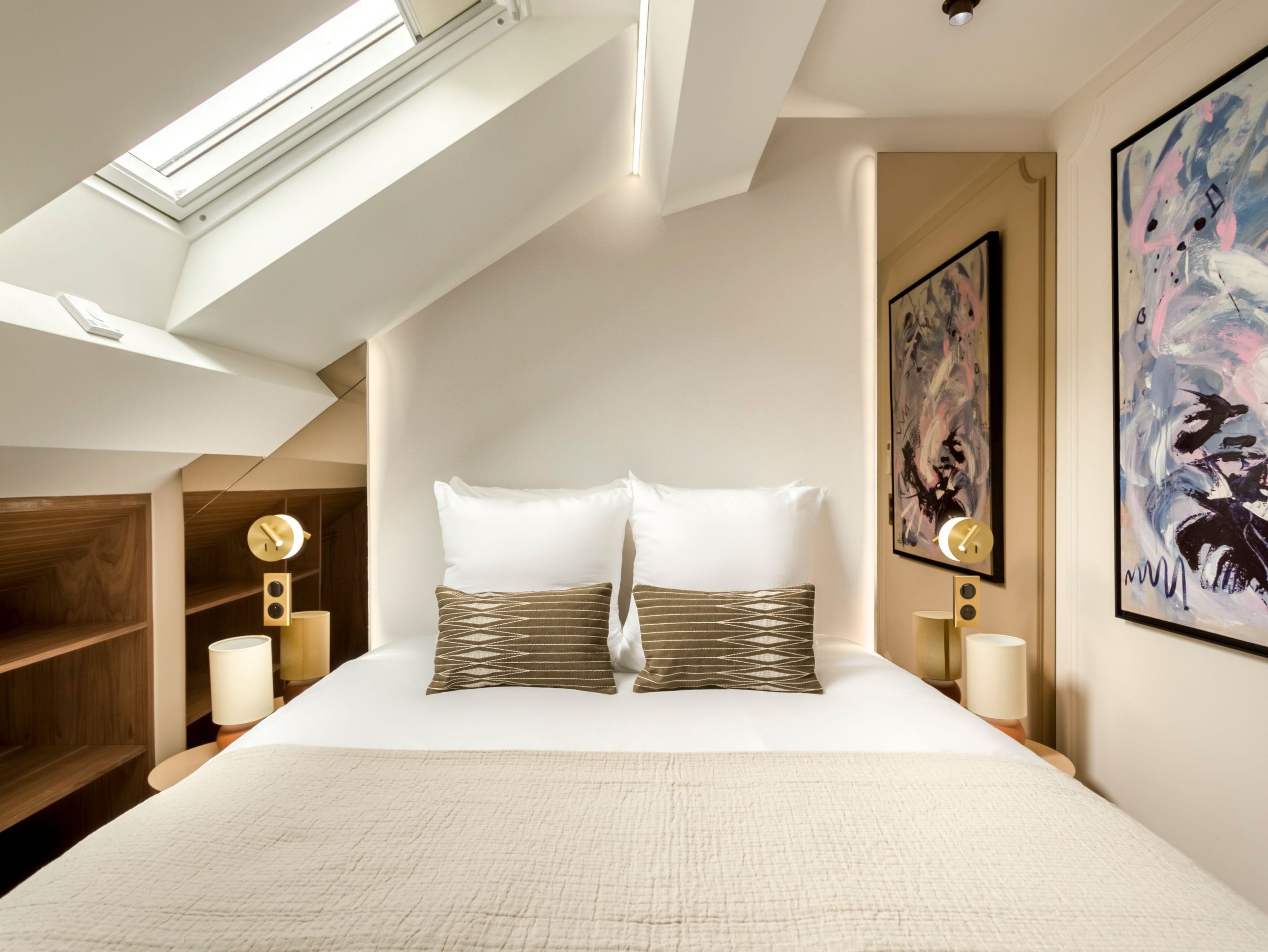 A modern bedroom with a skylight, a large bed with patterned pillows, two bedside lamps, and abstract wall art.