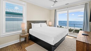 A bedroom with a large bed, a ceiling fan, a nightstand with a lamp, and a view of the ocean through a sliding glass door. - Cape San Blas Beach House - Starfish (Port St. Joe)