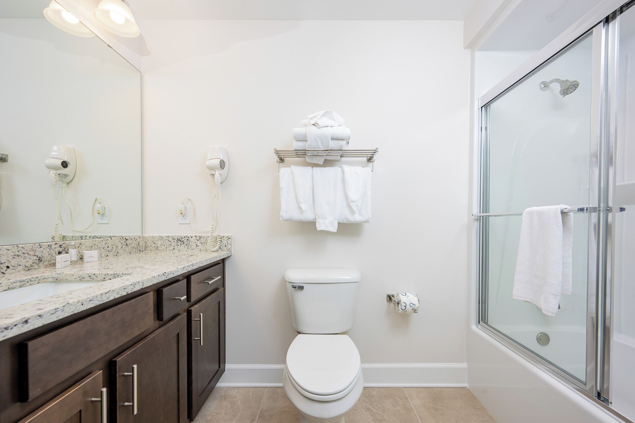 A bathroom with a toilet, a glass shower enclosure, a vanity with a marble countertop, and a towel rack with folded towels.