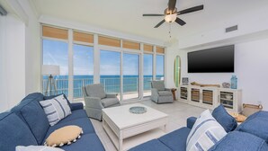A spacious living room with a large window overlooking the sea, a ceiling fan, and a flat-screen TV mounted on the wall. - Stunning Gulf views! - Phoenix Gulf Towers 1404 (Orange Beach)