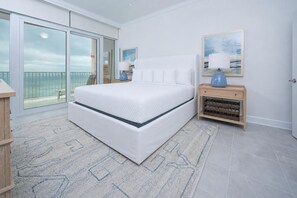 Un dormitorio con una cama grande, una mesita de noche, una lámpara y vistas al océano a través de una puerta corrediza.