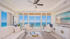 A spacious living room with a large window overlooking the ocean, a ceiling fan, and a flat-screen TV on a white console.