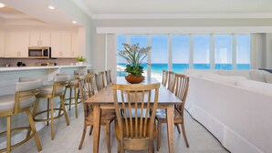 A modern kitchen with white cabinets, a dining area with wooden chairs, and a view of the ocean.