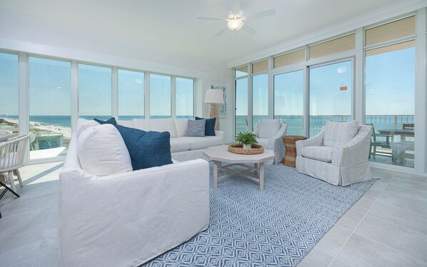 A spacious living area with a large window overlooking the beach, a ceiling fan, and a patterned rug.