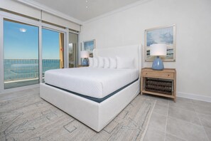 A bedroom with a large bed, a nightstand, a lamp, a framed picture, and a view of the sky through a sliding door.