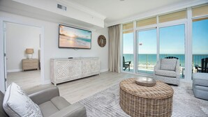 Een ruime woonkamer met een groot raam met uitzicht op een strand, een comfortabele bank, een rieten pouf en een witte kast met decoratieve panelen.
