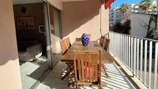 Un balcón con mesa y sillas de madera, un florero y un ventanal que da a un espacio interior.