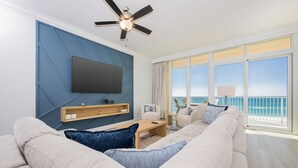 A modern living room with a large flat-screen TV, a ceiling fan, and a view of the ocean through large windows.