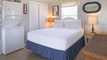 Un dormitorio con cama, lavadora, lámpara, silla de mimbre y espejo montado en la pared.