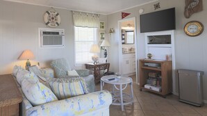 Una sala de estar acogedora con un sofá, un sillón y una pequeña mesa redonda. Se observa una estantería, un televisor de pantalla plana y un reloj en la pared.