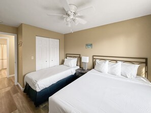Una habitación de hotel con dos camas individuales, un ventilador de techo, una mesita de noche con lámpara y una puerta que da a otra habitación.