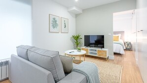 Un salon moderne avec un canapé gris, un téléviseur posé sur un meuble blanc et une plante.