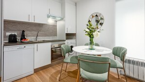 Une cuisine moderne avec des meubles blancs, un four encastré et une machine à café. On y trouve également un coin repas avec une table ronde et des chaises vertes.