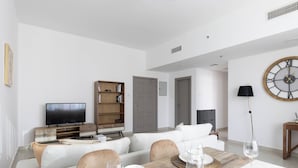 A modern living room with a sofa set, a dining table with placemats, a television, a bookshelf, and a wall clock. - Handsome 1BR in Armada Tower 3 (Dubai)