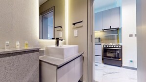 A modern bathroom with a white sink, mirror, and countertop. There are toiletries on the counter. The bathroom leads to a kitchen with a stove and oven. - Mag Eye 920 - Classy Studio Apartment in Meydan (Meydan)