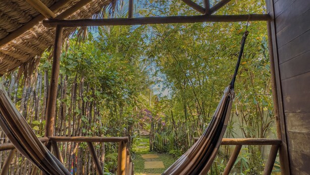 A hammock under a thatched roof structure with wooden benches and a view of a garden path. - Pousada Castelo do Vento (Cruz)