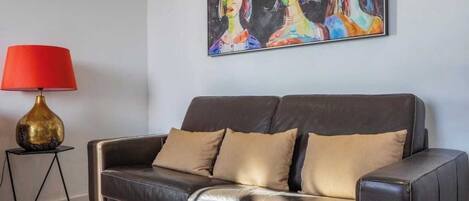 A living room with a dark leather sofa, a red lamp on a black metal stand, and a colorful painting on the wall.