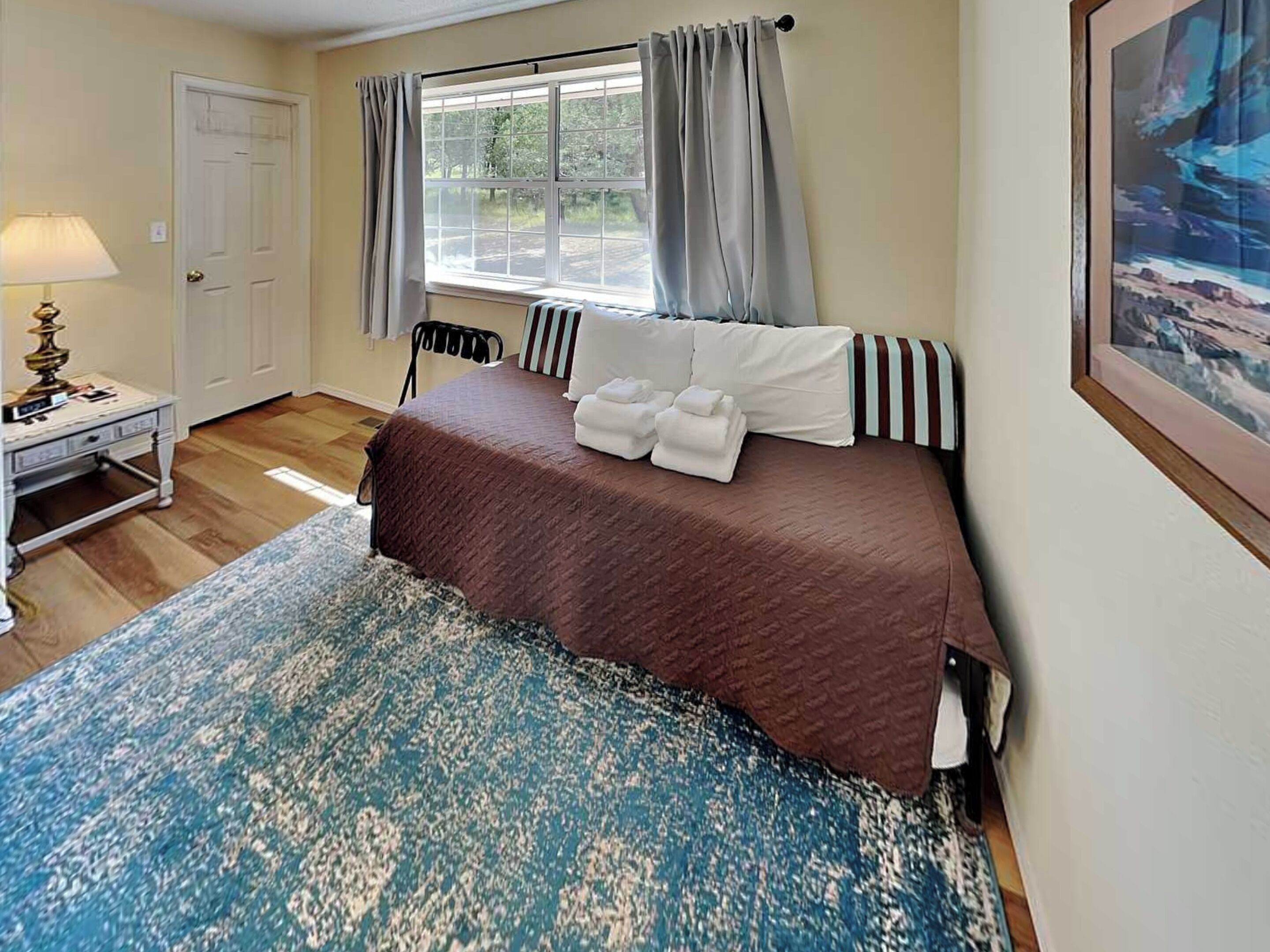 A hotel room with a bed, a desk, a window with curtains, and a painting on the wall.